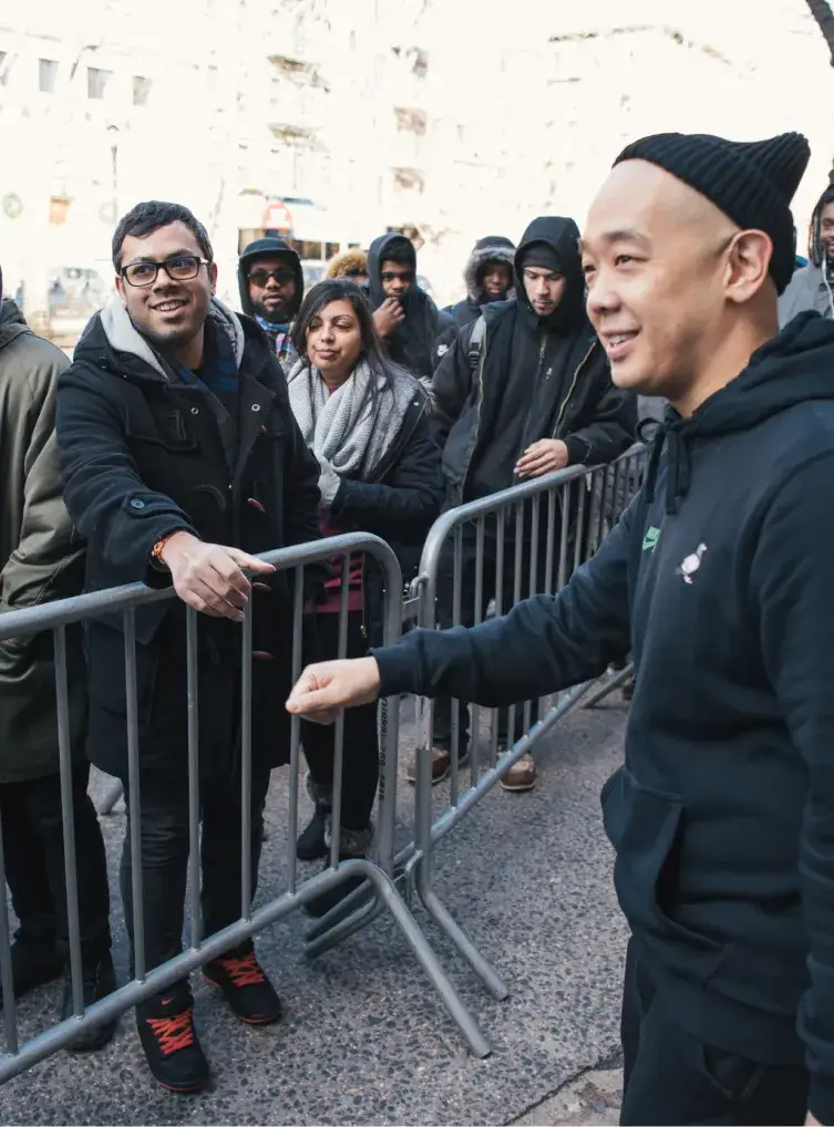 Man in a black hoodie and beanie greeting a man standing in a queue behind the fence.